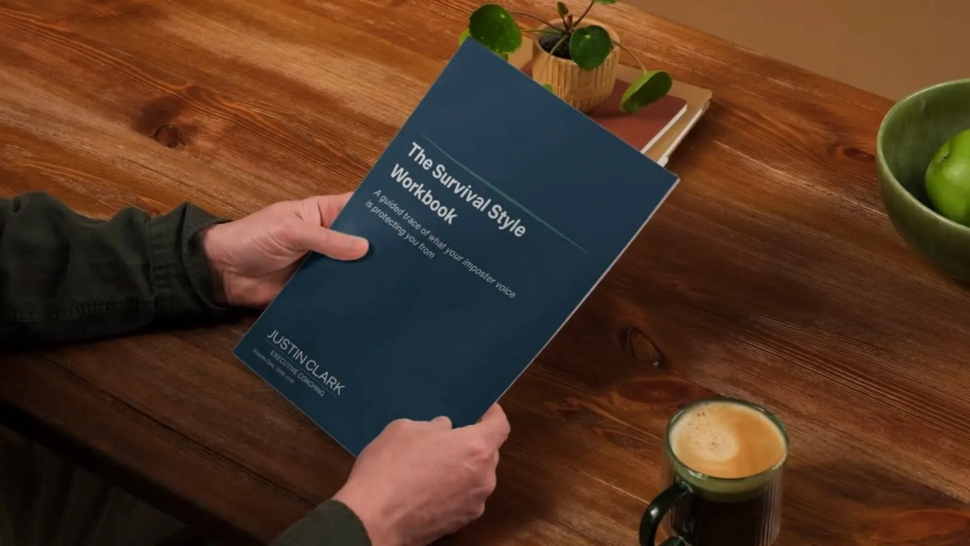 Person holding The Survival Style Workbook at a wooden table with a potted plant, a bowl of apples and a glass of coffee